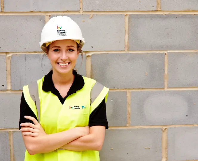 Female builder in front of wall with hard hat and hi-vis vest smiling into the camera